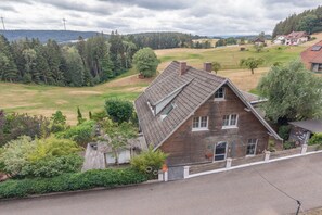 A two-story wooden house with a sloped roof, surrounded by a well-maintained garden and a rural landscape.