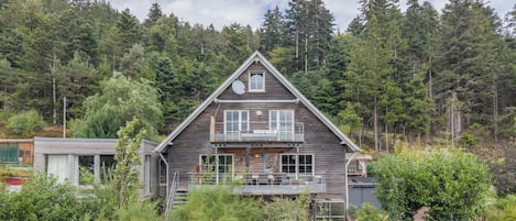 A two-story wooden house with a balcony, surrounded by dense forest.