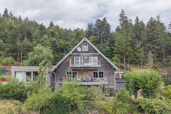 A two-story wooden house with a balcony, surrounded by dense forest.