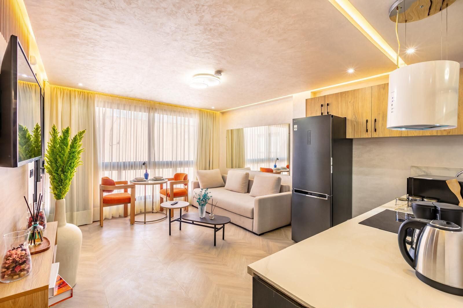Ein modernes Wohnzimmer mit Sofa, Couchtisch und Essbereich mit Stühlen. Es gibt eine Küche mit Kühlschrank, Herd und Wasserkocher.