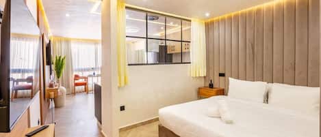 A modern hotel room with a large bed, a TV, a desk, and a dining area.