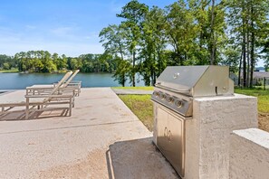 A barbecue grill with a concrete base, a set of wooden lounge chairs, and a view of a lake. - Lake Sinclair Dream Home w/ Everything|Pool|HotTub (Milledgeville)