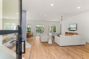 Un salon spacieux doté d’un plancher en bois, de canapés blancs, d’un coin repas avec une table et des chaises, et d’une grande fenêtre offrant une vue sur la verdure.