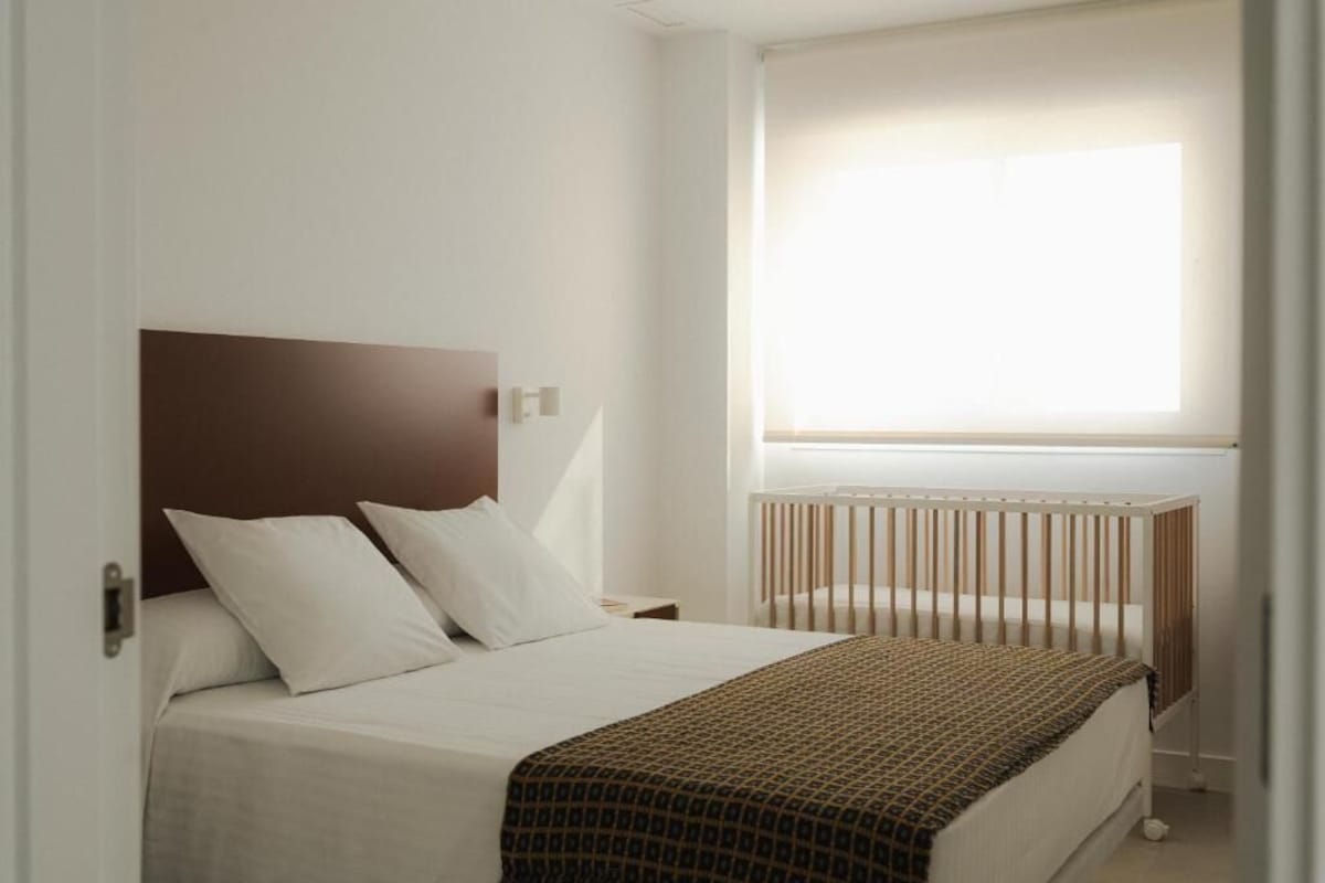 Ein Schlafzimmer mit einem Bett, einem Kopfteil, einem Fenster mit Jalousien und einem Babybett.