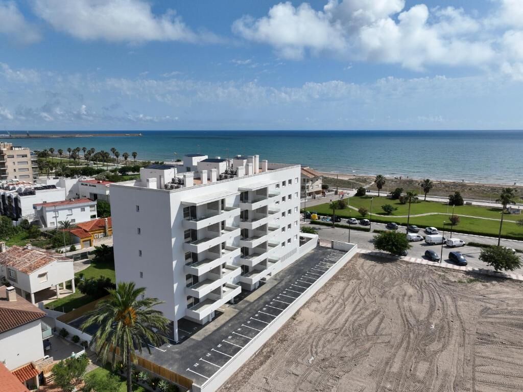 A multi-story building with a parking lot in front, adjacent to a sandy beach and the sea.
