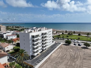 Ein mehrstöckiges Gebäude mit Parkplatz vor der Tür, direkt neben einem Sandstrand und dem Meer.
