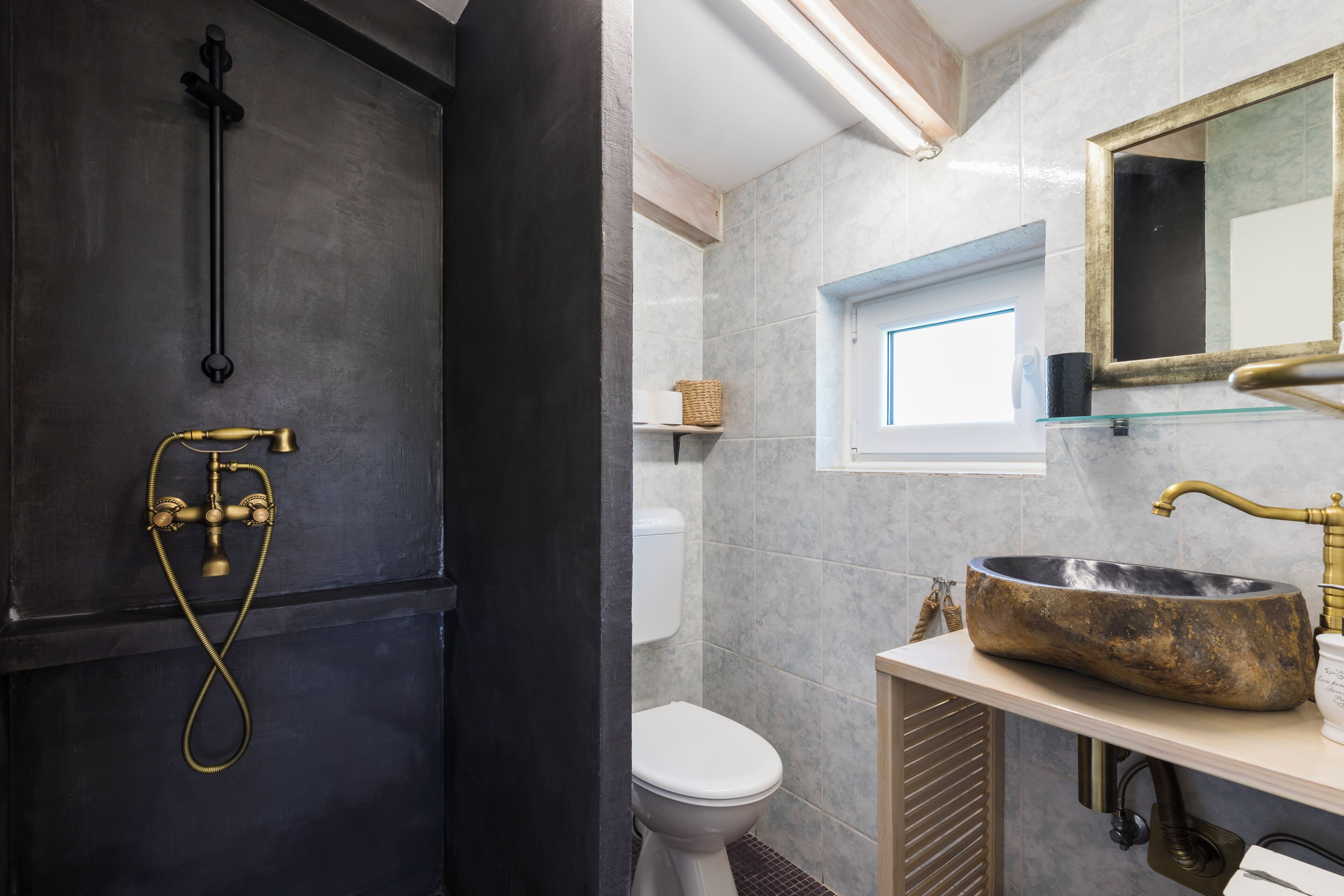 Baño con pared negra, grifería de ducha de latón, inodoro blanco y un lavabo de piedra grande con un espejo encima.