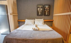 Un lit avec du linge de lit blanc et une serviette enroulée, une tête de lit en bois et deux éléments décoratifs encadrés accrochés au mur.