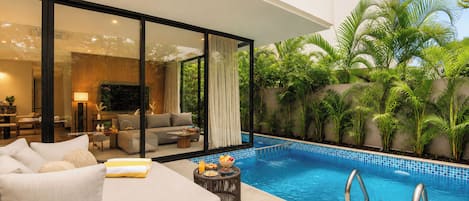 A modern outdoor pool area with a sofa and dining table, surrounded by lush greenery and a clear blue pool.