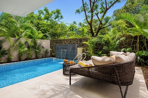 A poolside area with a wicker sofa, a small table with a bowl of fruit and glasses, and a stone wall.