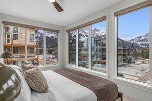 A bedroom with a large bed, a ceiling fan, and a view of a mountain landscape through large windows. - Modern 4BR|Hot Tub and Mountain Views|Steps from Downtown (Canmore)