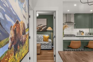 A modern kitchen with green cabinets, a dining area with a wooden table, and a living room with a sofa and a painting. - Modern 4BR|Hot Tub and Mountain Views|Steps from Downtown (Canmore)