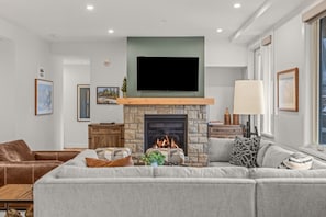 A modern living room with a stone fireplace, a flat-screen TV mounted above it, and a large grey sofa. - Modern 4BR|Hot Tub and Mountain Views|Steps from Downtown (Canmore)