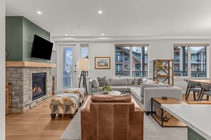 A modern living room with a stone fireplace, a grey sectional sofa, and a flat-screen TV mounted on the wall. - Modern 4BR|Hot Tub and Mountain Views|Steps from Downtown (Canmore)