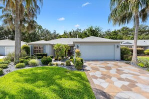 A residential property with a well-maintained lawn, a driveway leading to a garage, and a variety of landscaping including shrubs and palm trees. - Oasis in the Sun - tropical retreat w/ 4-seater golf cart (The Villages)