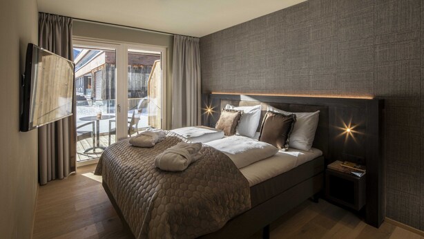 A modern hotel room with a large bed, a flat-screen TV mounted on the wall, and a view of the outdoor area through a sliding glass door. - Apartment mit 2 Schlafzimmern (Westendorf)