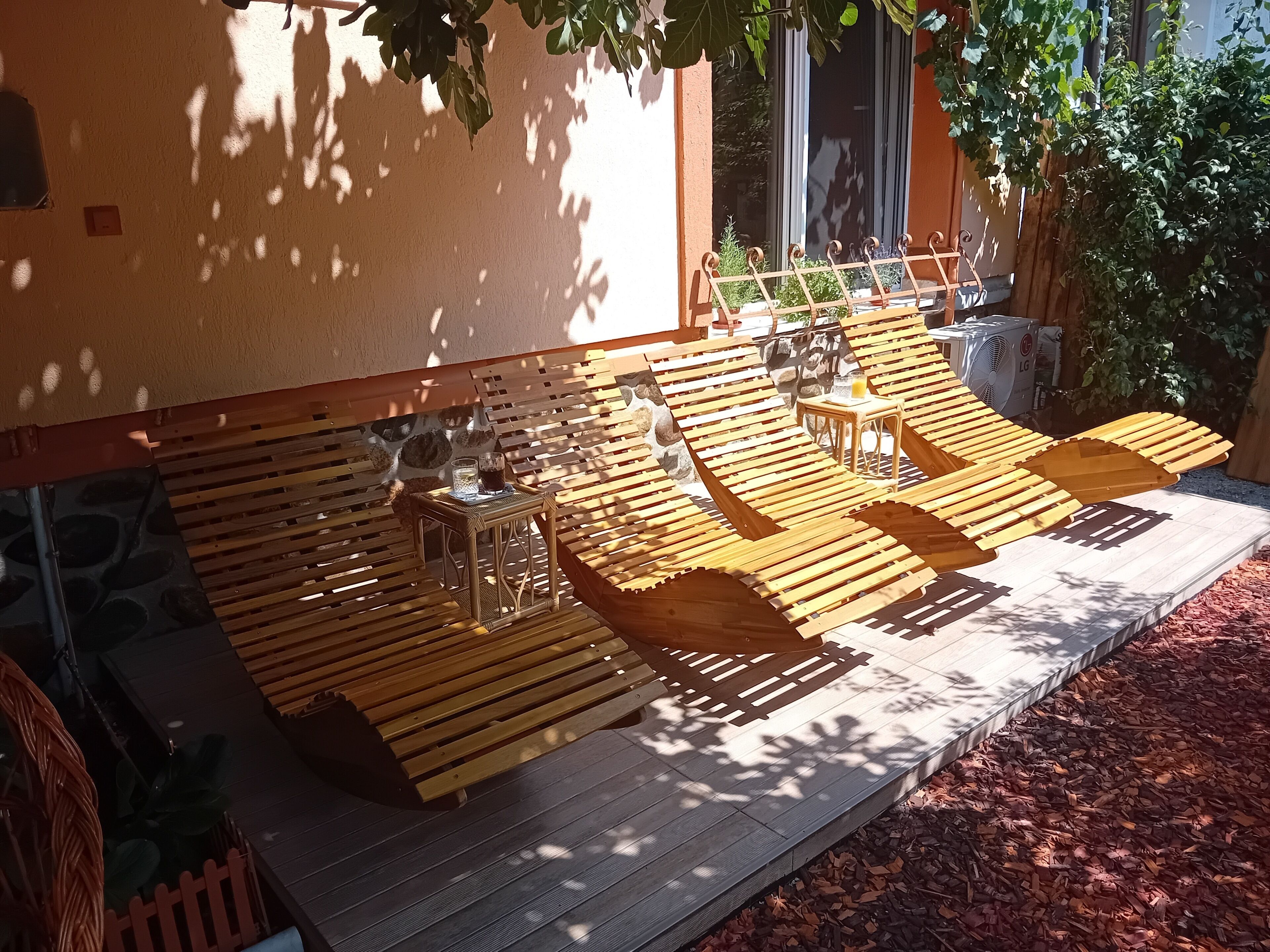 Un espace extérieur aménagé avec des chaises longues en bois et une petite table, entouré d’un mur de pierre et de verdure.