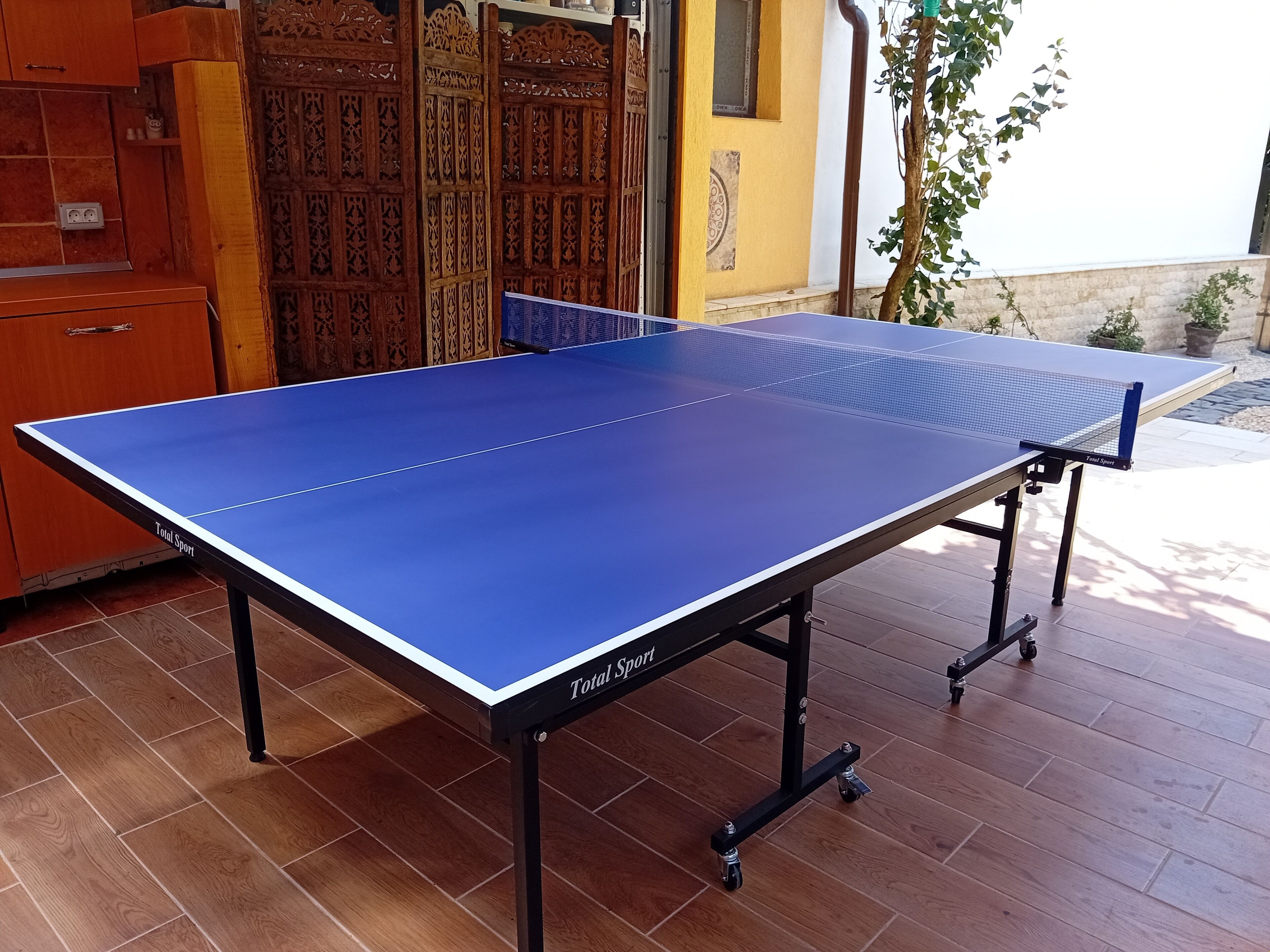 A blue table tennis table with 'Total Sport' branding, placed on a wooden floor with a cabinet and decorative wooden panels in the background.