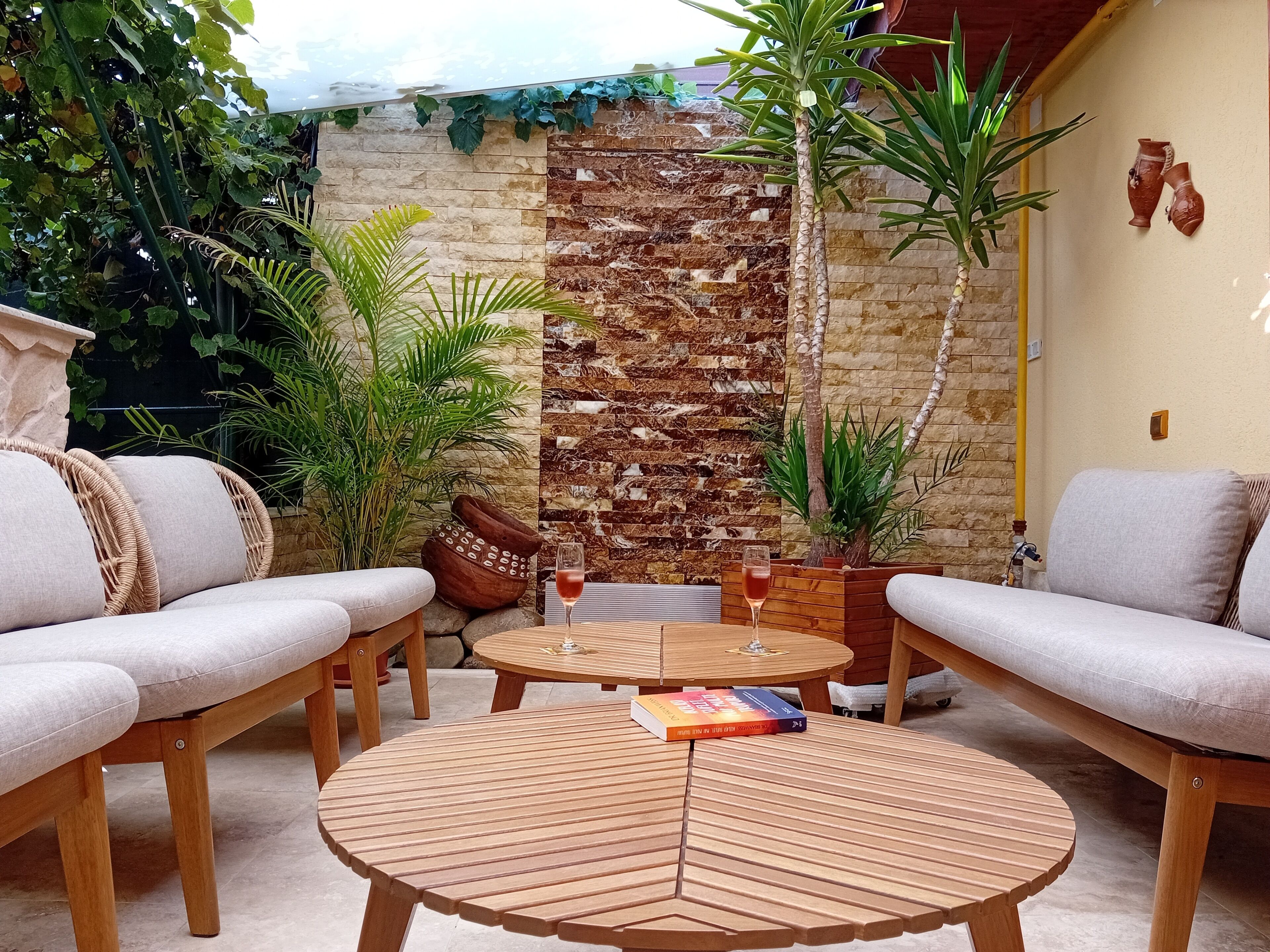 Un rincón exterior acogedor con una mesa redonda de madera, sofás con cojines y varias plantas.