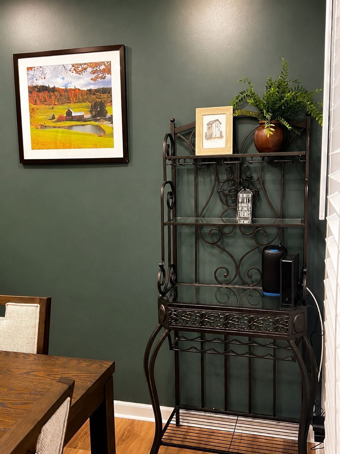 A room with a dining table, a framed picture on the wall, a metal shelf with a plant and a small framed picture, and a black speaker on the shelf.