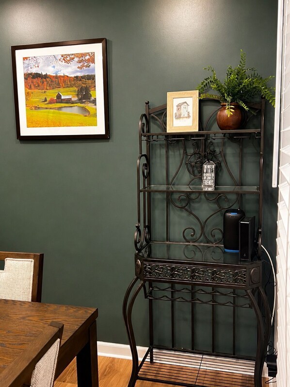 A room with a dining table, a framed picture on the wall, a metal shelf with a plant and a small framed picture, and a black speaker on the shelf.