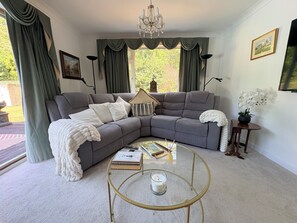 A living room with a grey sectional sofa, a round glass coffee table, a chandelier, and a view of greenery outside.