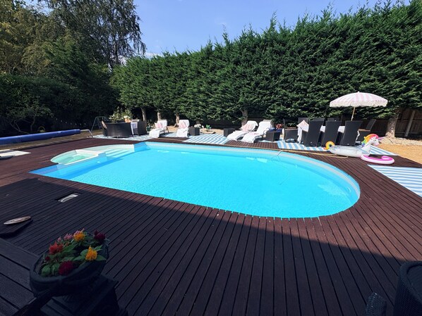 A swimming pool with lounge chairs and umbrellas, surrounded by a wooden deck and a hedge.