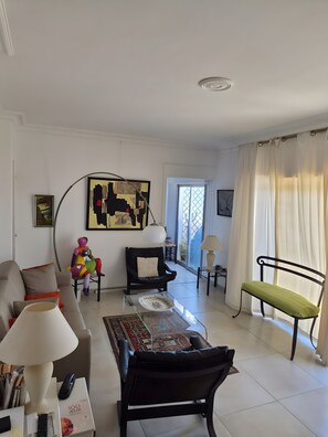 Ein Wohnzimmer mit einer Couch, einem Sessel, einem Tisch und einer Lampe. Ein Fenster mit Vorhängen und ein Gemälde an der Wand.