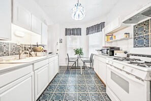 A kitchen with white cabinets, a tiled floor, a dining table with chairs, a stove, and a hanging light fixture. - Seward St - Rooms - Shared Bath & Spaces (Rochester)