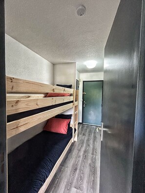 Una cama litera con estructura de madera y una cama individual en la parte inferior. Se observa una puerta al fondo.