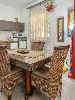 Un comedor con una mesa redonda, sillas de mimbre, una cocina moderna al fondo y un cuadro abstracto colorido en la pared.