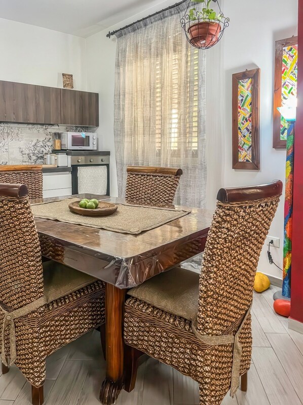 Un coin repas avec une table ronde, des chaises en osier, une cuisine moderne en arrière-plan et un tableau abstrait aux couleurs vives accroché au mur.