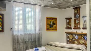 Ein Hotelzimmer mit einem Bett, einem Gemälde, einem an der Wand befestigten Regal mit Tellern und einem Fenster mit durchsichtigen Vorhängen.