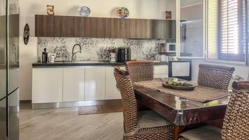 A modern kitchen with a dining area, featuring a wooden table, wicker chairs, and a large window with blinds.