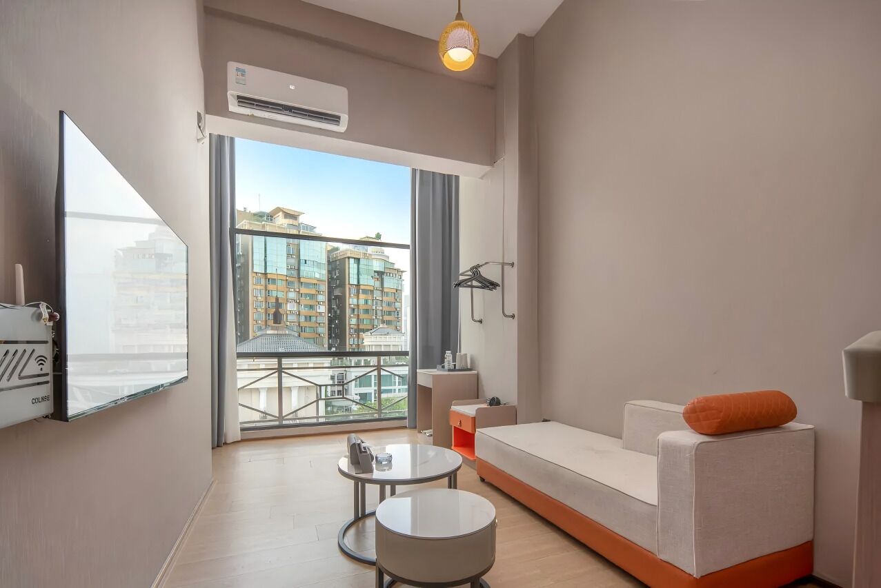 A modern hotel room with a sofa, a small table, a TV, and a balcony with a view of buildings.