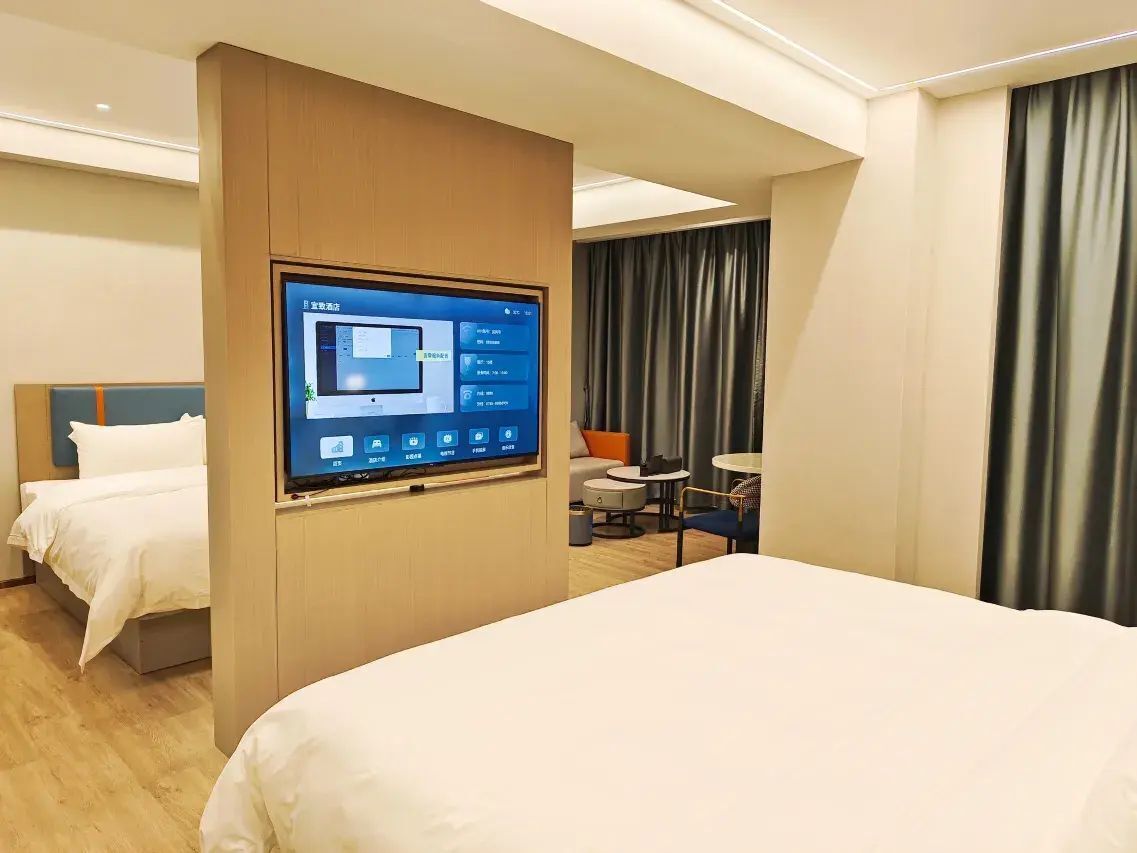 A hotel room with a bed, a TV mounted on a wooden panel, a desk with a chair, and a window with curtains.