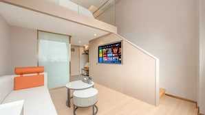 A modern hotel room with a sofa, a small table, a chair, and a TV mounted on the wall displaying a menu.