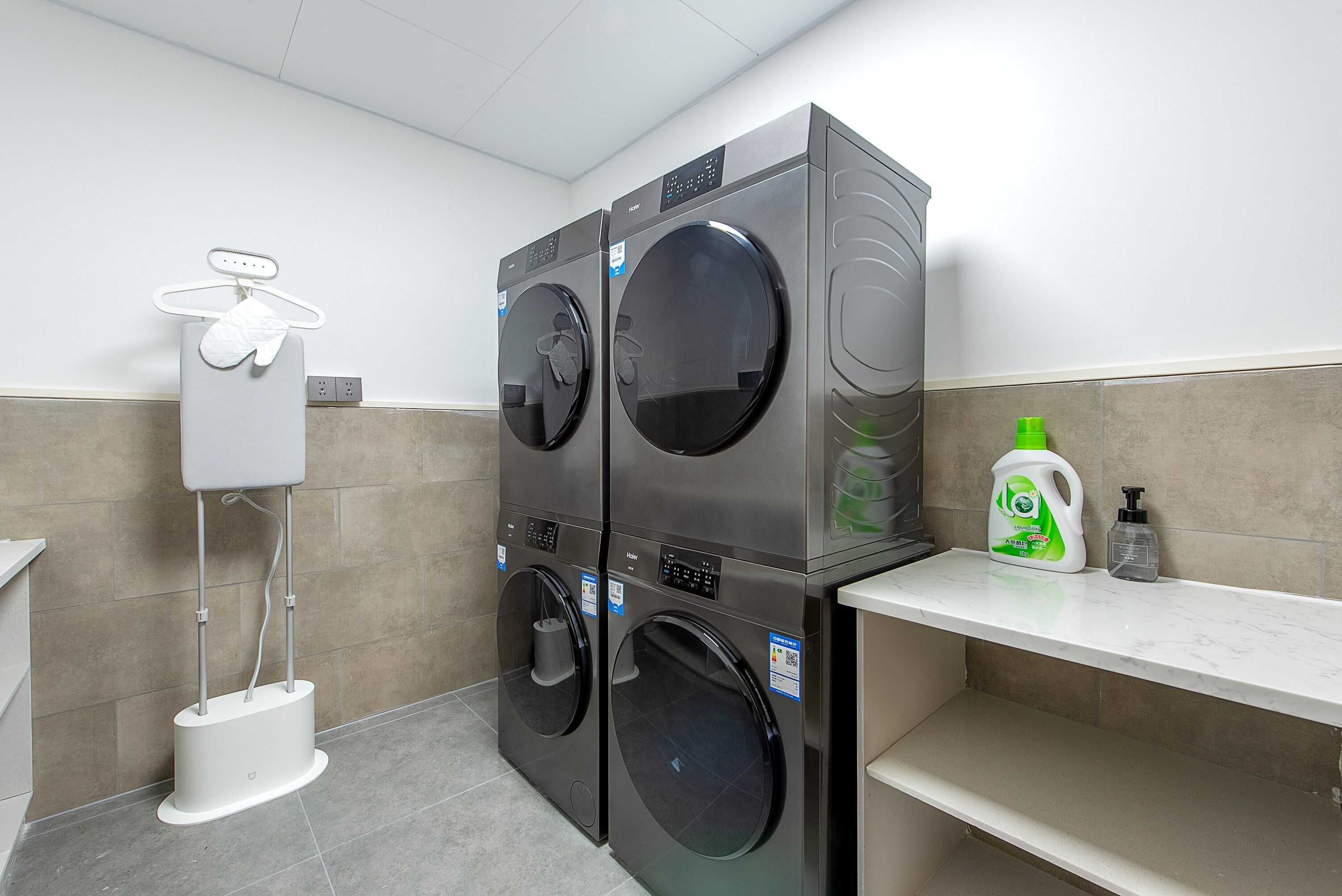 A laundry facility with two stacked washing machines, a drying machine, a white utility box, and a shelf with cleaning supplies.