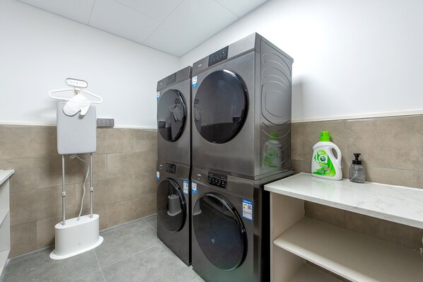 A laundry facility with two stacked washing machines, a drying machine, a white utility box, and a shelf with cleaning supplies.