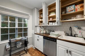 A kitchen with white cabinets, a dishwasher, a window with a view of trees, and a countertop with various kitchen items. - Historic cottage-Same builder as Grove Park Inn-HOT TUB-15 mins to ASHEVILLE (Swannanoa)