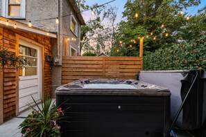 Un jacuzzi en un patio trasero con valla de madera, luces de cadena y una casa de fachada de piedra.