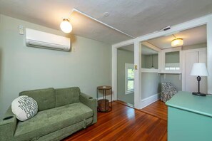 A room with a green sofa, a small round table, an air conditioning unit, a lamp, and a door leading to another room. - Historic cottage-Same builder as Grove Park Inn-HOT TUB-15 mins to ASHEVILLE (Swannanoa)