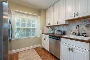 Una cocina con armarios blancos, lavavajillas, ventana con persianas y salpicadero de azulejos.