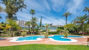 A pool area with a clear sky, surrounded by palm trees and a well-maintained lawn. - 1 Dormitorio en Medina Garden Ref 712 (Marbella)