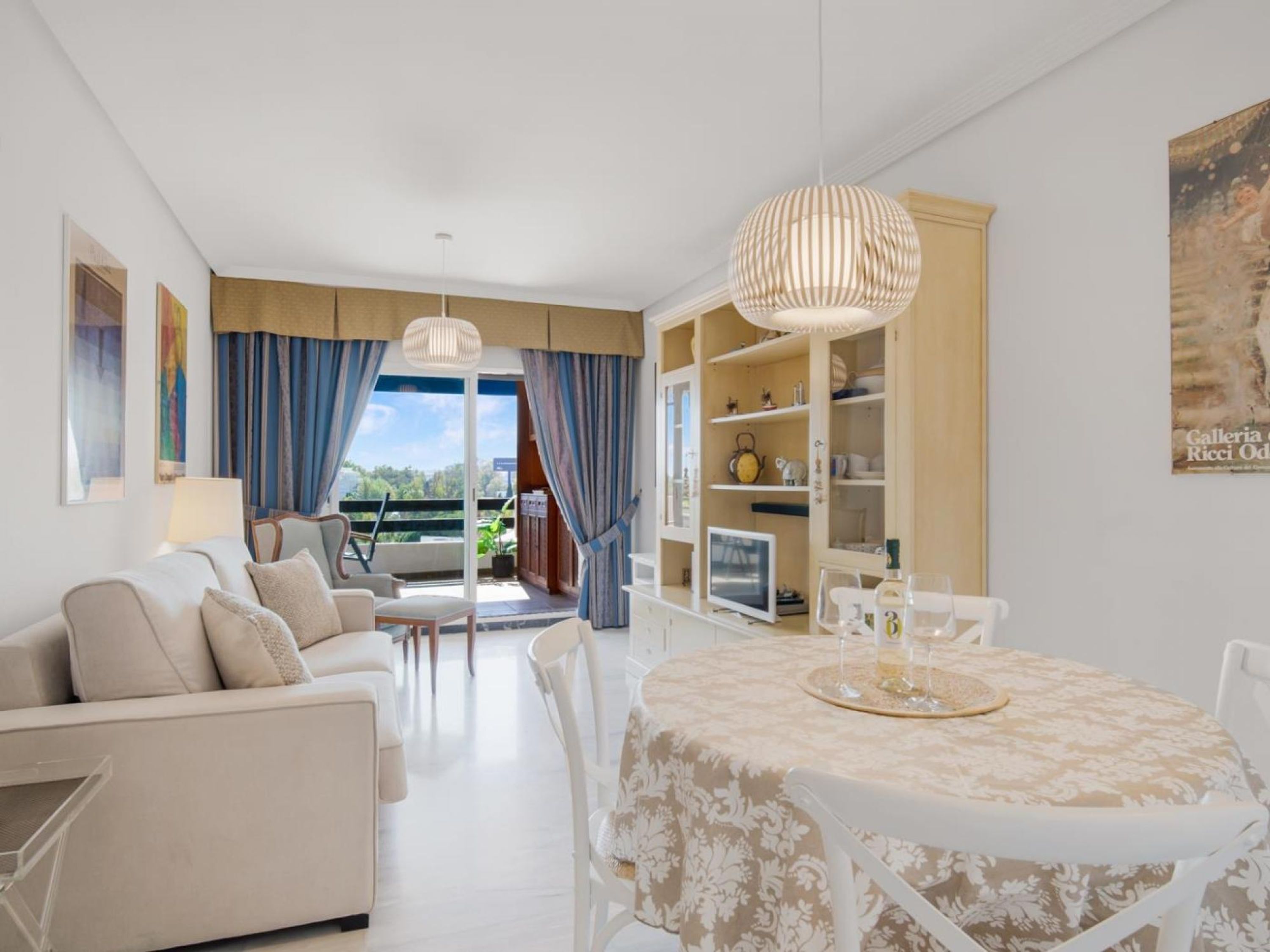 Una sala moderna con sofá, sillas, una mesa de comedor con mantel estampado y vistas a un área exterior a través de un ventanal.