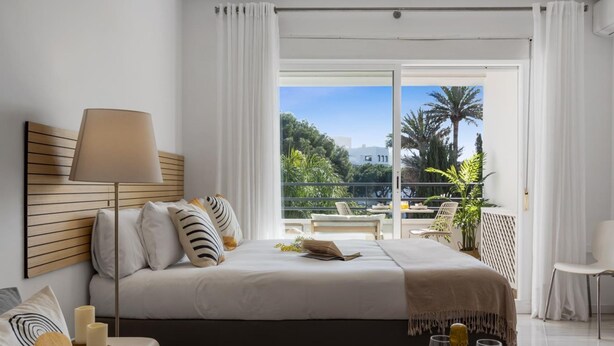 A modern bedroom with a large bed, a bedside table with candles, a lamp, and a view of a balcony with outdoor furniture. - Acogedor Estudio en Medina Garden ref 122 (Marbella)