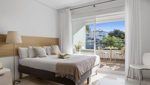 A modern bedroom with a large bed, a bedside table, a lamp, and a view of a balcony with outdoor furniture. - Acogedor Estudio en Medina Garden ref 122 (Marbella)