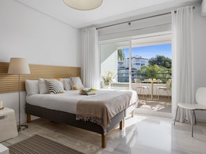 A modern bedroom with a large bed, a bedside table, a lamp, and a view of a balcony with outdoor furniture. - Acogedor Estudio en Medina Garden ref 122 (Marbella)