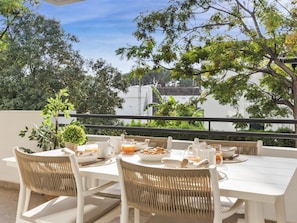 An outdoor dining area with a white table and wicker chairs, overlooking a garden and buildings. - 2 Dormitorios en Medina Garden Ref 511 (Marbella)