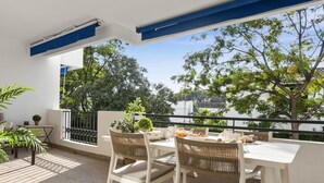 Ein Balkon mit weißem Tisch und Stühlen, einem kleinen Blumentopf und Blick auf Bäume und Gebäude.